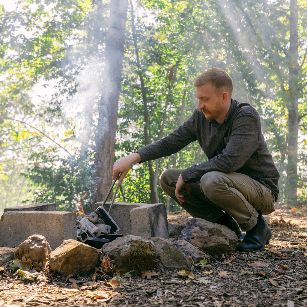 Teamaker crouching beside a charcoal fire in the woods arranging charcoal to stoke fire.