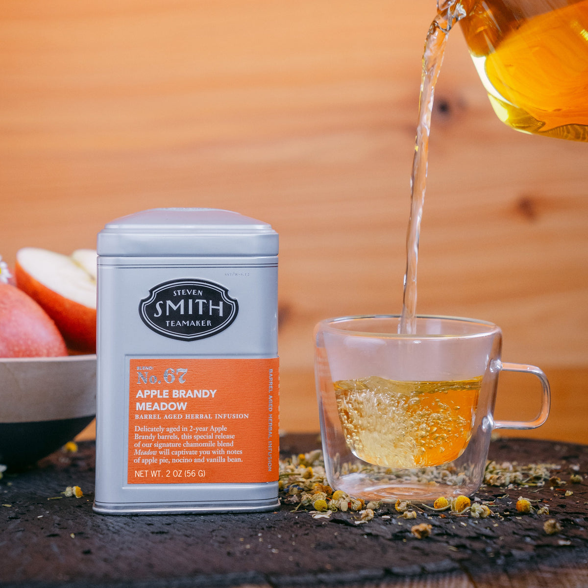 Grey tin of Apple Brandy Meadow with an orange label next to a teacup being filled with brewed tea and a bowl of apples in the background.
